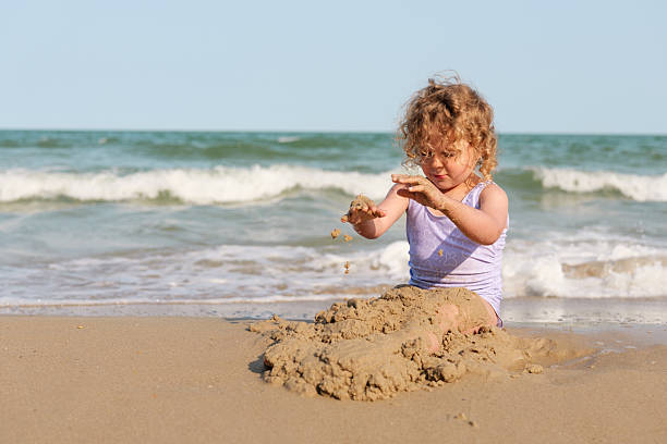 istock A young girl sits on the sandy shore, focused on building a sandcastle while waves wash gently nearby under a clear blue sky 2238122700