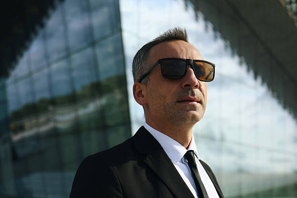 Caucasian middle aged smartly dressed man wearing sunglasses standing outdoors in front of modern glass building looking confidently into distance, business professional posing for corporate concept