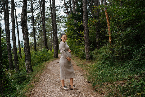 Side view of a pregnant woman in a beige knit dress and sneakers, standing on a gravel forest path. She gently cradles her baby bump with one hand while looking at the camera. Tall pine trees and a wooden railing create a peaceful natural backdrop.