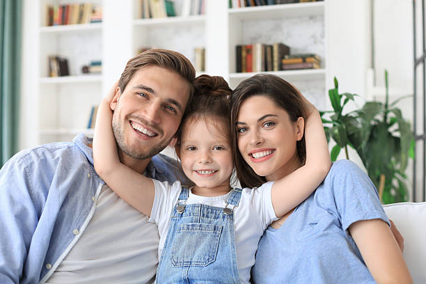 istock Positive friendly young parents with smiling little daughter sitting on sofa together while relaxing at home on weekend. 2238126855