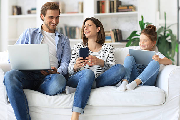 istock Father, mother and daughter using electronic devices sitting on sofa at living room. 2238127029