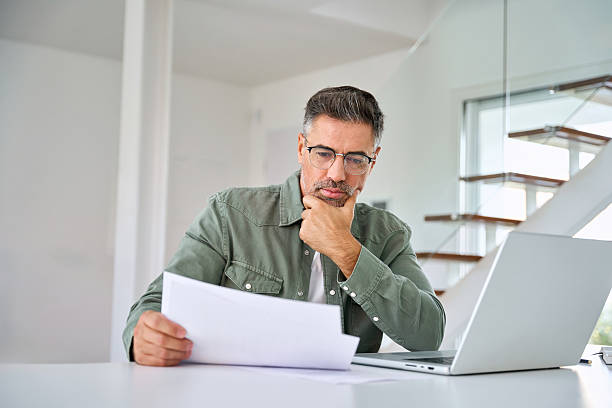 Serious mature man looking at paper bill working at home using laptop computer.