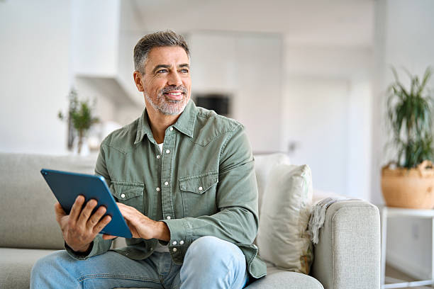 istock Happy mature man using digital tablet relaxing on couch looking away at home. 2238131379