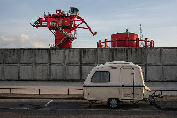 A small vintage caravan parked in front of a tall concrete wall in an industrial zone of Rotterdam, the Netherlands. Bright red industrial machinery and storage tanks rise above the wall, creating a stark contrast between leisure and heavy industry. The scene is captured in soft evening light, highlighting the juxtaposition of domestic travel and urban infrastructure.
