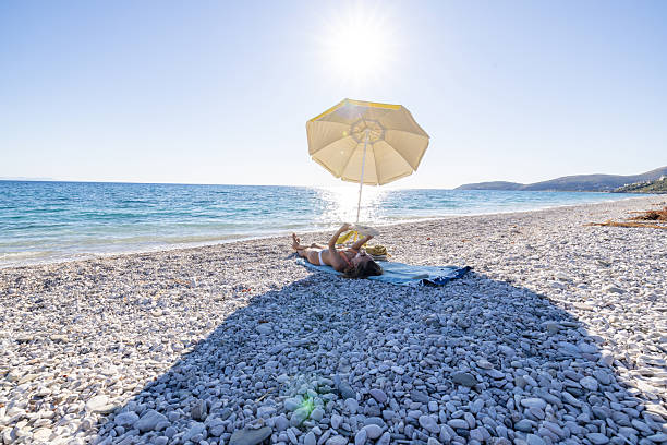 Young woman in bikini lying on blue mat under yellow umbrella on rocky beach. Relaxing by the sea on a sunny day with clear blue sky. Summer vacation and leisure time at the coast.