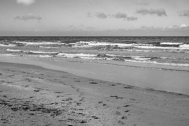 Soft waves roll onto a sandy beach under a cloudy sky. The shoreline shows scattered footprints leading towards the water, creating a tranquil, inviting scene.