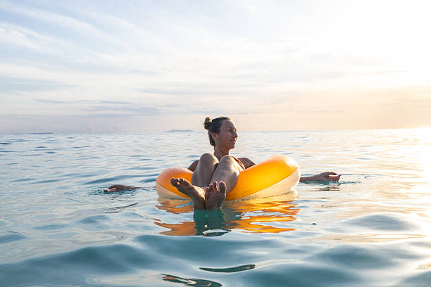 Smiling woman in swimsuit lounging on yellow inflatable ring in calm ocean water during sunset. Leisure and relaxation on water. Adult enjoying peaceful beach vacation moment.