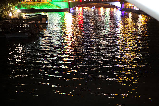 A photograph of a nighttime cityscape with colorful lights reflecting on the rivers surface. In the distance, a bridge and illuminated embankments create a picturesque atmosphere of the urban night scenery