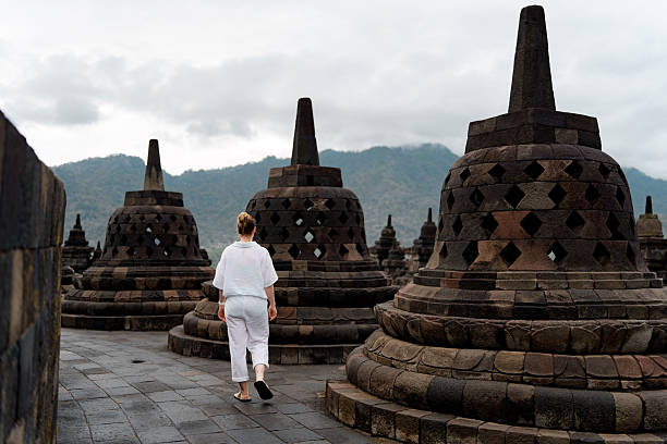 A woman walking among ancient stone stupas at a historical site, with mountains in the background and a cloudy sky