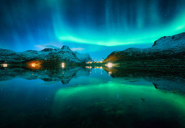 The aurora borealis illuminates the night starry sky, bathing snowy mountains in a green glow and reflected on a tranquil lake.  Winter wonderland in Lofoten Islands, Norway.  Northern lights. Space