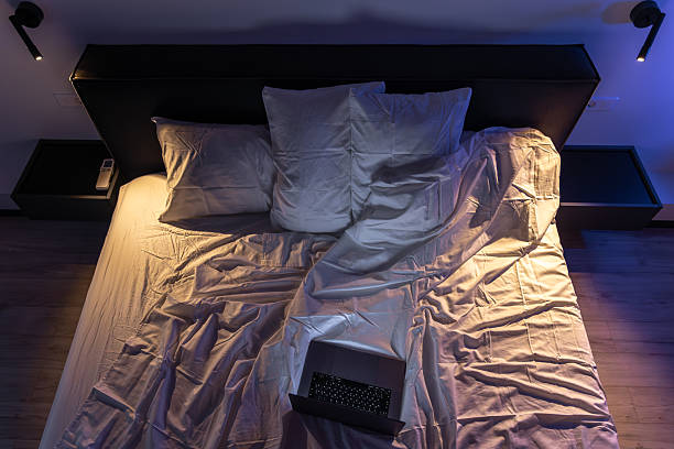 Overhead shot of a bed with a lit keyboard laptop, strong contrast of blue and warm light, pronounced bedding folds, a quiet atmosphere for late night work and focus.