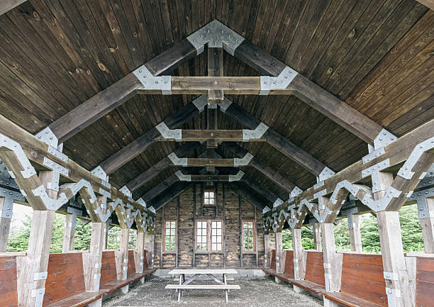 A spacious rustic wooden shelter with exposed timber trusses, metal brackets, and long benches along both sides. Natural light filters through windows, creating a warm, nostalgic setting ideal for outdoor gatherings and picnics.