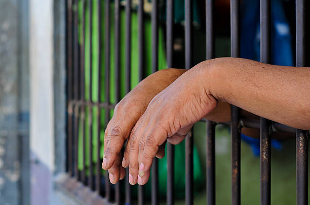 istock prisoner's hands behind bars with black background 2238170785