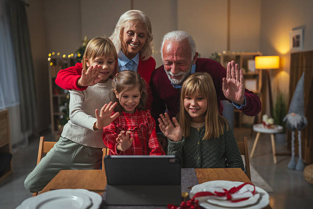 Family including grandparents and children waving while having a virtual video call communication on a tablet, celebrating connecting during christmas amidst festive decorations