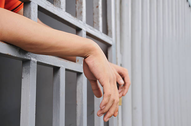 istock prisoner's hands behind bars with black background 2238174632
