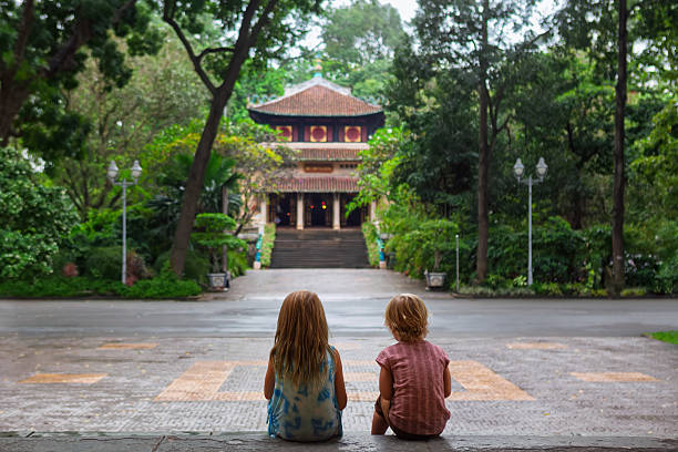 Two children sit on steps facing a traditional Vietnamese temple surrounded by lush tropical greenery in Saigon. Peaceful moment in cultural setting, perfect for family travel, and lifestyle themes.
