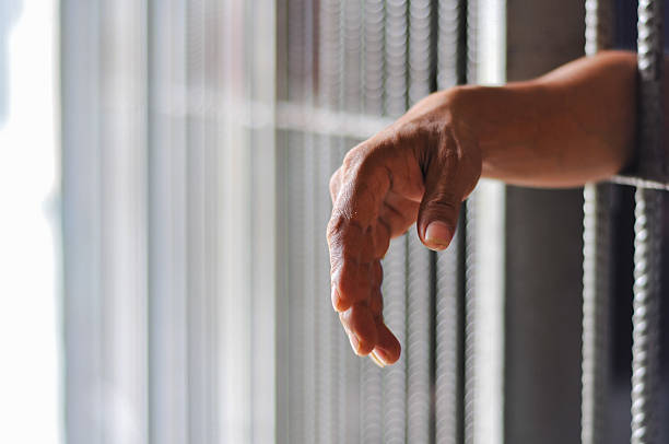istock prisoner's hands behind bars with black background 2238174726