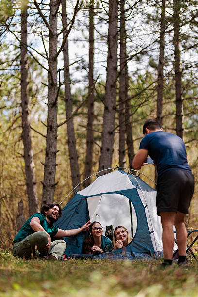 istock Friends enjoying a fun camping trip while setting up a tent in a serene forest, surrounded by tall trees on a sunny day 2238188149
