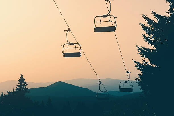 Chairlift gliding over coniferous trees with majestic mountains rising in the background, bathed in the warm hues of sunset