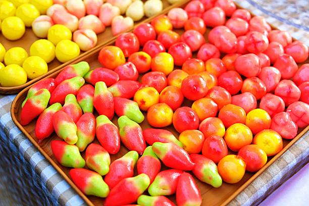 Close up of colorful Thai Look Chup desserts shaped like fruits, made from mung beans and coconut milk, displayed neatly on a wooden tray.