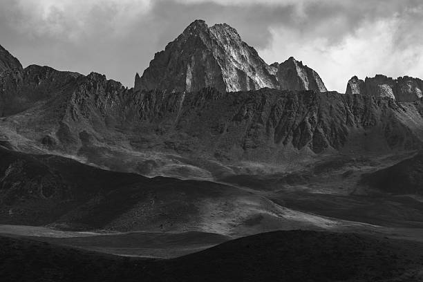 This striking monochromatic landscape captures the raw majesty of towering, sharply ridged mountain summits piercing a brooding sky filled with swirling dark clouds, with layered rugged foothills descending into shadowed valleys below. The high-contrast textures and ethereal light evoke isolation, power, and untamed wilderness, ideal for adventure travel, nature inspiration, mountaineering visuals, or atmospheric editorial in advertising, posters, or digital media projects.