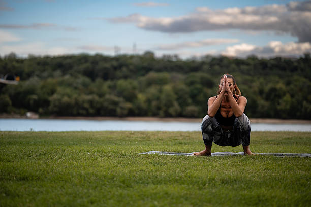 Yoga instructor practicing garland pose on a mat in a serene park by a lake, surrounded by lush greenery and a cloudy sky