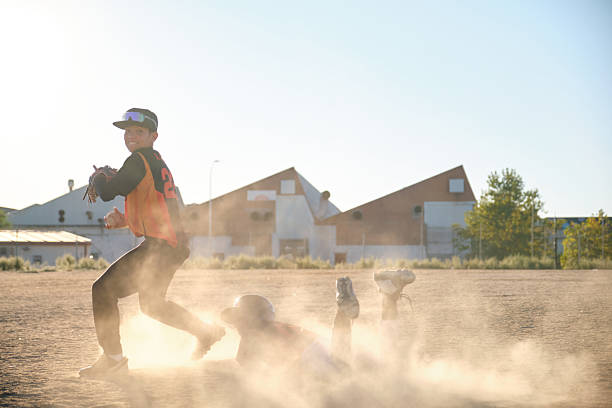 Boy sliding into base while teammate fields ball in cloud of dust