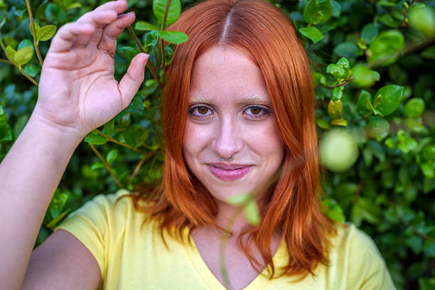 Young woman with red hair and septum piercing smiling, bleach eyebrows, enjoying nature with green leaves