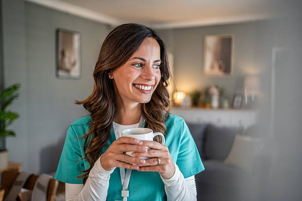Mid-30s woman nurse in scrubs smiles while holding a mug and relaxing during a peaceful coffee break at home, enjoying a quiet moment of self-care by the window
