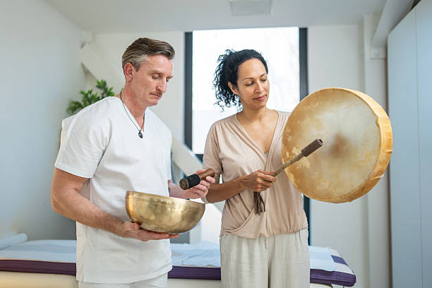 Mid-age Caucasian male therapist and Hispanic female therapist performing sound therapy indoors using a golden bowl and drum in a bright, serene setting.