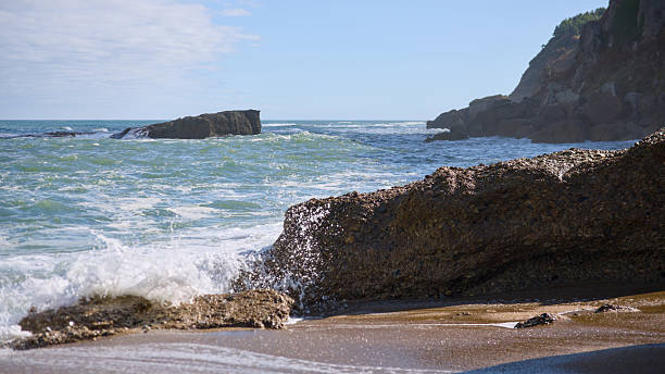 Ocean waves breaking against rocks on a sandy beach with clear sky and scenic cliffs in the background. Natural seascape, travel and ecology concept.
