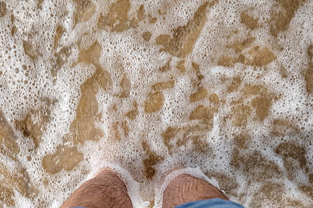 Caucasian male bare feet and pulled up jeans, standing on a sandy beach with shallow tidal waves sea water reaching the shore. Top down, close up view