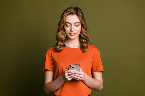 istock Smiling young woman with blond curls using a smartphone against a khaki background in casual orange attire 2238270020