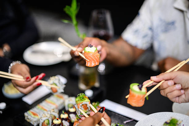 Close up of hands with chopsticks lifting salmon roses and avocado sushi from a shared platter at a restaurant table
