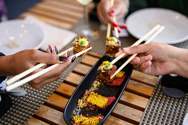 Closeup of hands with chopsticks reaching for crispy pork belly served with charred corn and ginger from an appetiser plate