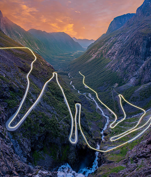 Woman wearing an orange jacket walks along a quiet fjord road surrounded by steep cliffs and autumn-colored hills in Norway.