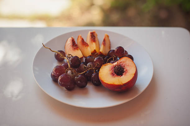 Horizontal close-up view of a halved peach with slices and grapes on a white plate, placed on a white wooden table. Floral shadows create a natural pattern in the bright midday Mediterranean light. Minimal composition with copy space, evoking freshness, simplicity, and summer atmosphere. Filmed in Sardinia, Italy.