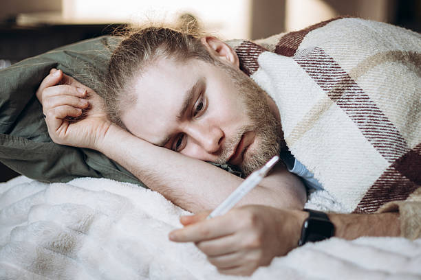 istock Man lying in bed at home, covered with a blanket, looking at a thermometer. 2238294471