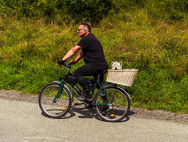 Czorsztyn, Poland – August 6, 2025: A man rides a green bicycle along a rural road with his small white dog sitting in a wicker basket. Dressed in black sportswear, he enjoys outdoor activity on a summer day.