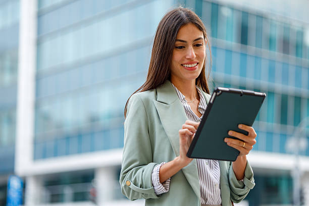 istock Businesswoman smiling while using a digital tablet outdoors, standing in front of a modern city building 2238303633