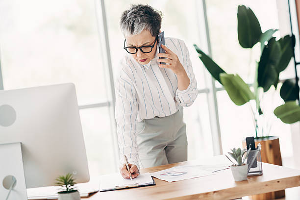 istock Mature businesswoman in a modern office working at a desk while multitasking, writing notes, and speaking on the phone 2238305146