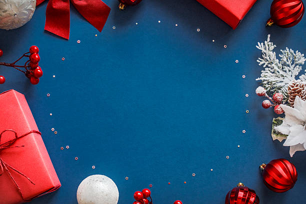 istock Festive Christmas frame with red baubles, gift boxes, berries, frosted pine branches, ribbon bows and confetti, trinkets, and white and red ornaments on dark blue background with copy space in the middle 2238318499