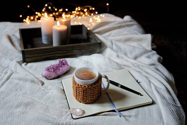 A mug of tea sits on a notebook near a crystal and pen. Candles in the background glow with fairy lights. The scene is set on a white blanketed bed, giving it a sense of reflection and peace. Copy space for text, high angle view - Alternative therapy, concepts and ideas