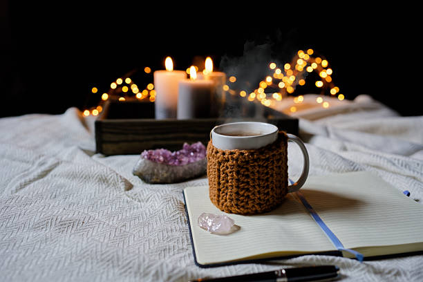 A mug of hot tea with a knitted mug cover sits on an open notebook next to amethyst and quartz crystals. Candles and fairy lights glow softly in the background. Copy space for text, high angle view - Spirituality, alternative therapy
