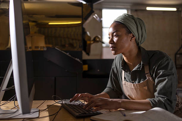 A woman wearing a head wrap and apron concentrates at a desktop computer in a workshop, illustrating focus, skill, and productive work in a hands-on environment