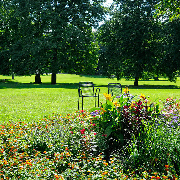 A lush green lawn and vibrant flower beds are featured in a tranquil park scene with two empty metal chairs. The image captures the serenity of nature and offers ample copy space for text.