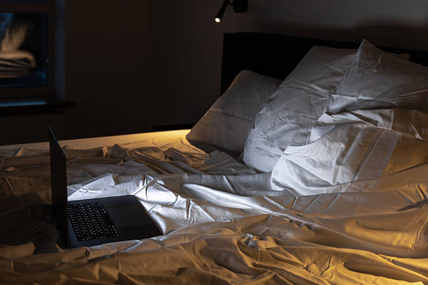 istock Side view laptop on rumpled bed under lamp. Late binge. 2238354833