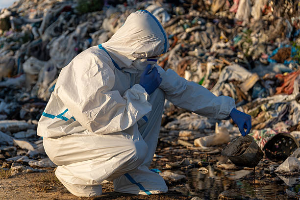 istock A worker in a protective suit crouches in front of a landfill, inspecting waste while the sun sets in the background, focusing on pollution 2238360039