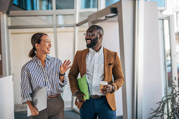 Two diverse business colleagues walk and chat through a modern office, smiling and carrying a laptop, documents, and coffee as they discuss work and collaborate confidently