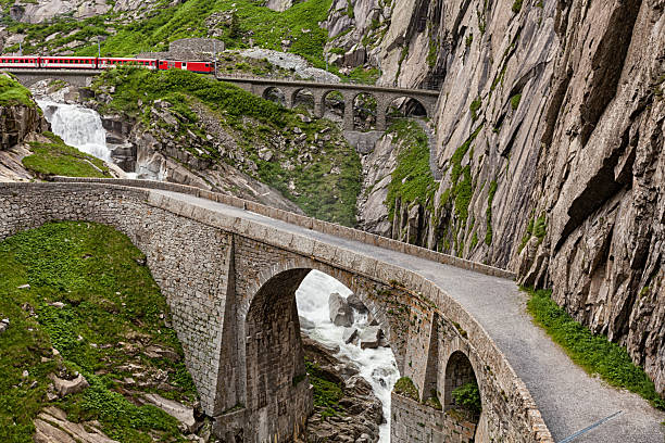 Red train on historic Devil’s Bridge above waterfall in Schollenen Gorge, Andermatt, Switzerland, surrounded by rocky cliffs.